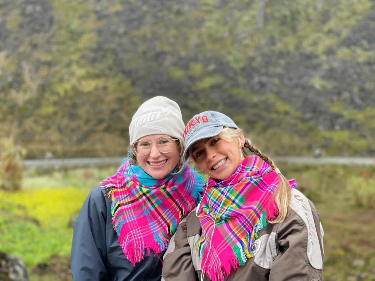 Two smiling women wearing colorful scarves and hats pose outdoors with a blurred green landscape in the background. One hat reads "Tokyo".