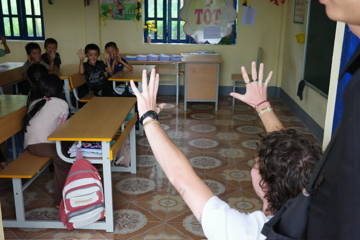 Person playfully raises hands in a classroom with smiling children seated at desks. "TỐT" poster on the wall. Warm, cheerful atmosphere.
