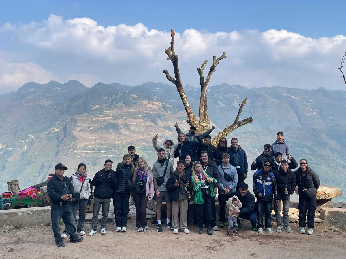 Group of people posing happily on a mountain with a scenic view. A leafless tree is in the background. Clear skies and clouds above.