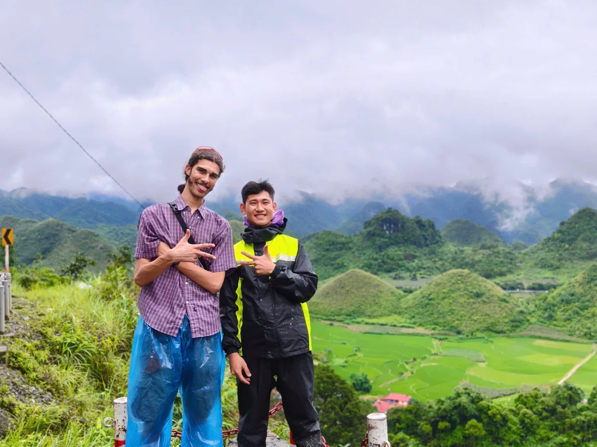 Two smiling people pose with peace signs in front of a lush, green mountain landscape under cloudy skies. One wears a bright jacket.