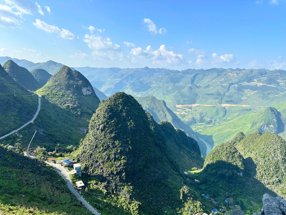 Lush green mountains under a clear blue sky with scattered clouds. A winding road runs through the landscape, creating a serene, vast scenery.