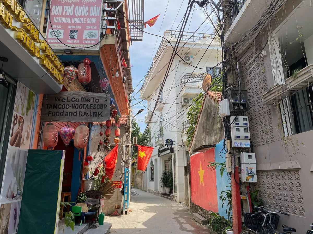 Narrow alley with colorful lanterns, Vietnamese flags, signs for noodle soup, and tangled wires. Bright, sunny day.