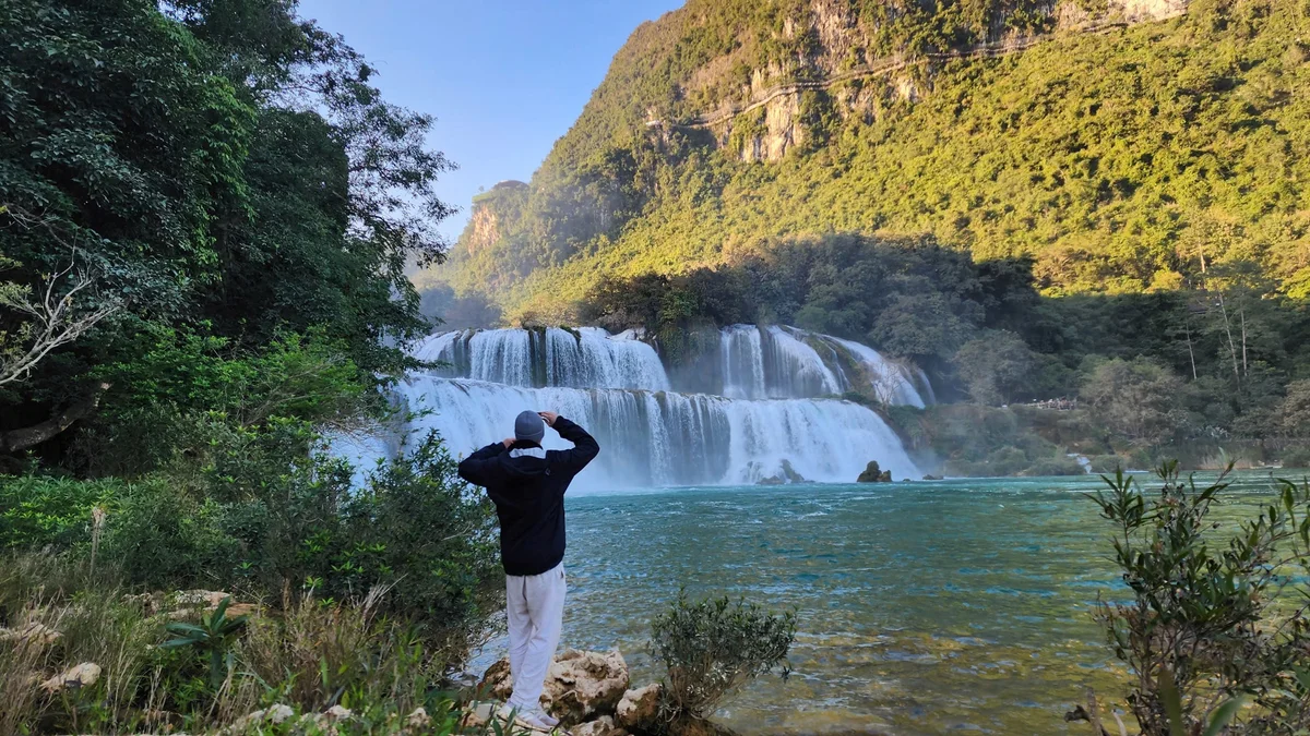 Person in a black jacket and white pants views a large waterfall in a lush, green mountainous landscape. Clear blue water in the foreground.