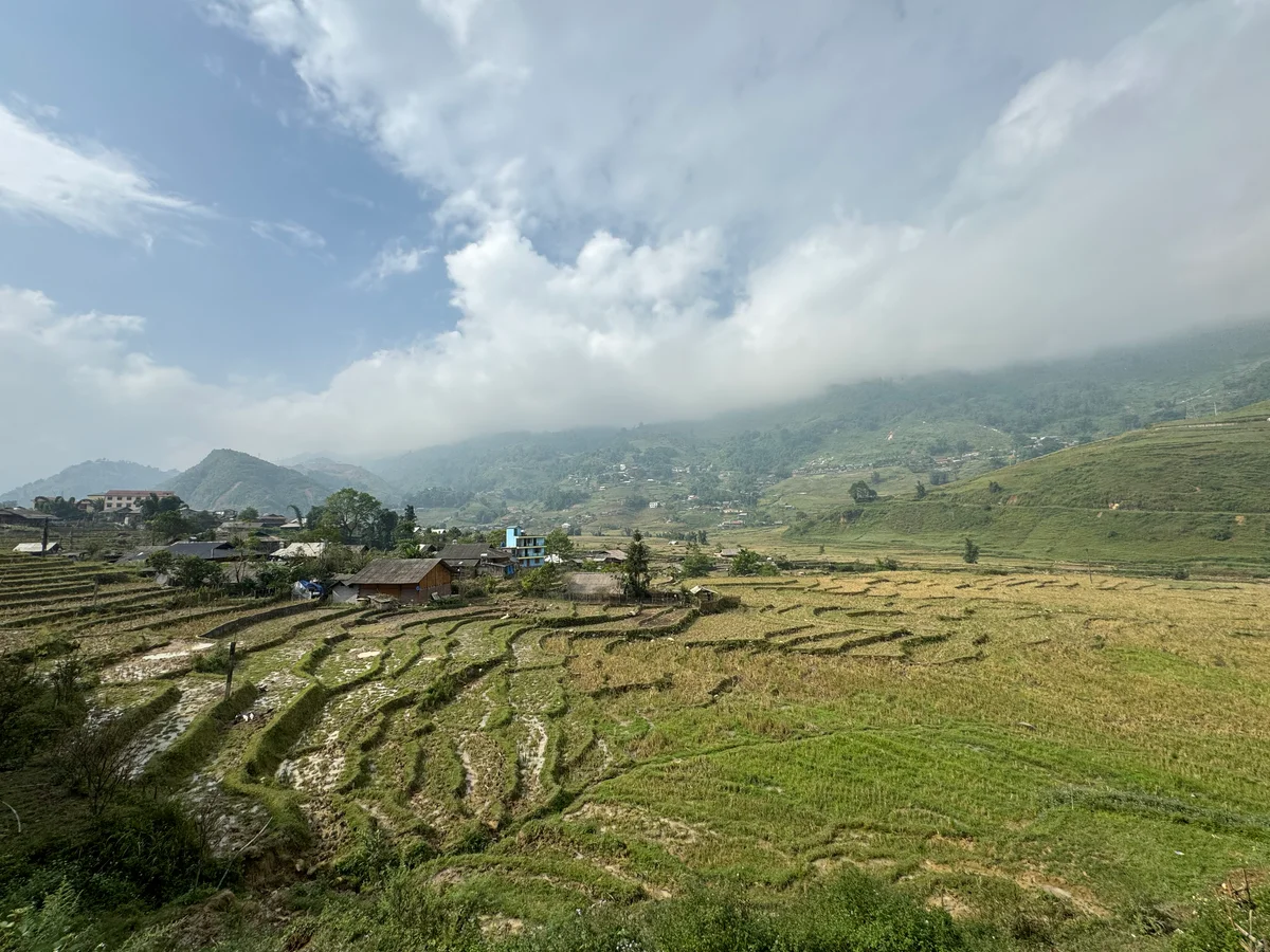 Terraced fields under a cloudy sky with mountains in the background. A small village with several houses is nestled among the greenery.