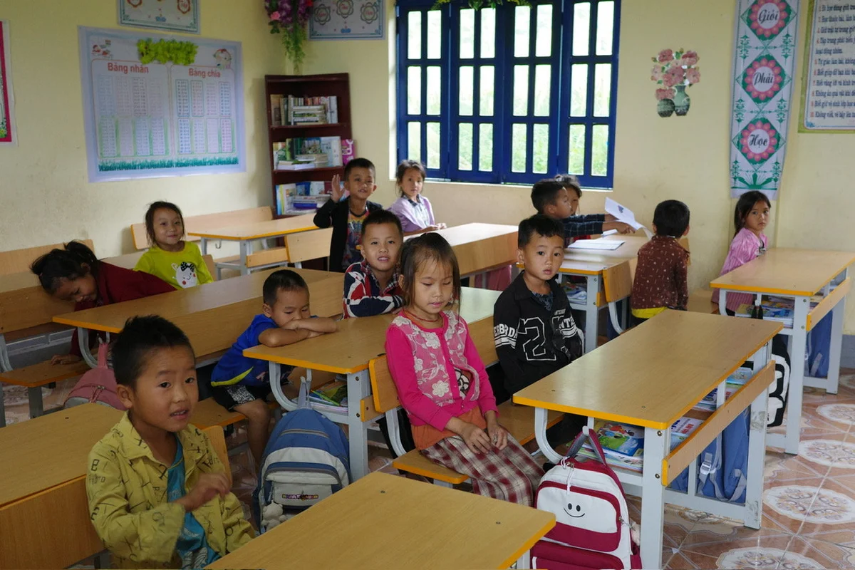Children sit at desks in a colorful classroom, some smiling, with backpacks on tables. Posters and a window view are in the background.