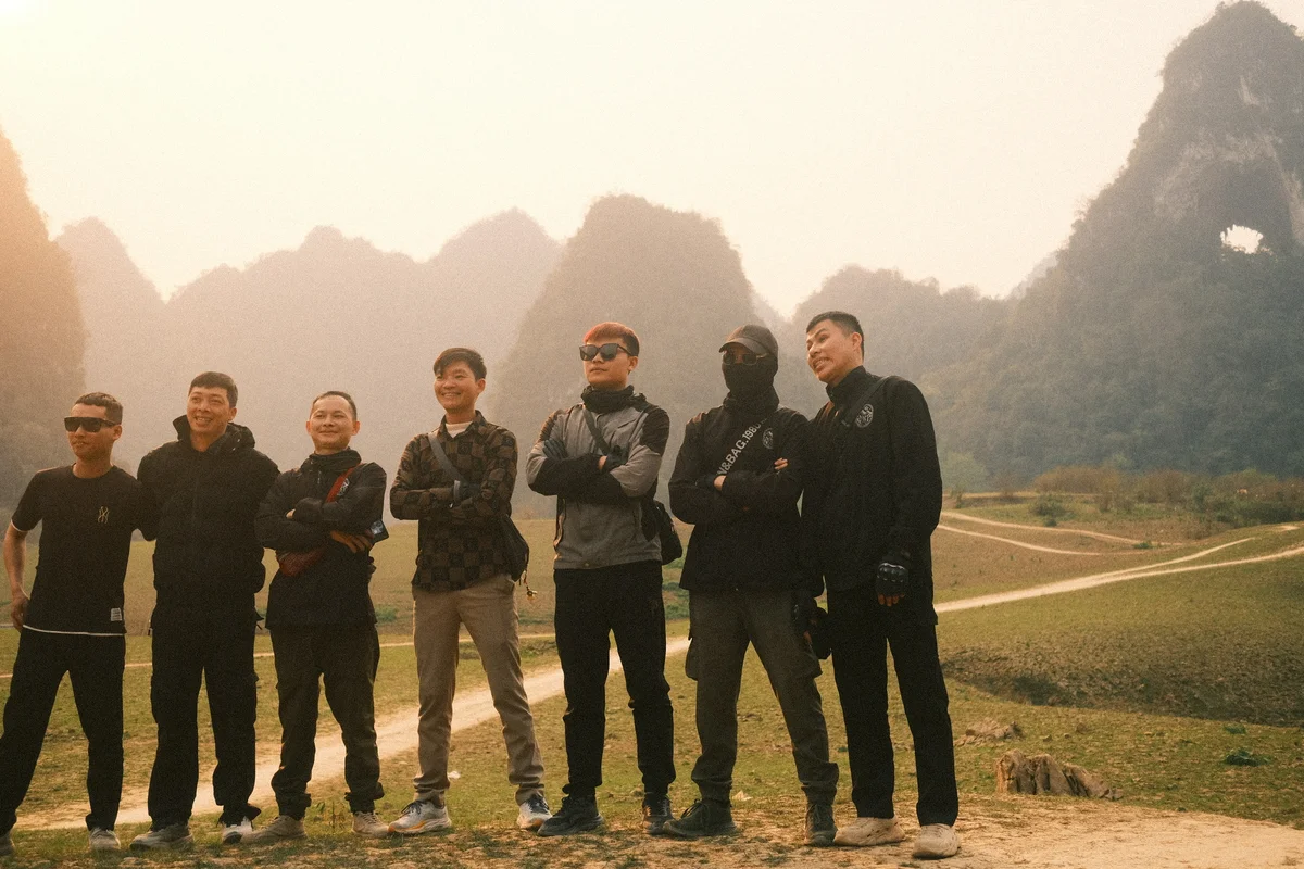 Seven men pose smiling outdoors with arms crossed against a misty mountain backdrop. Casual attire in earthy tones conveys a relaxed mood.