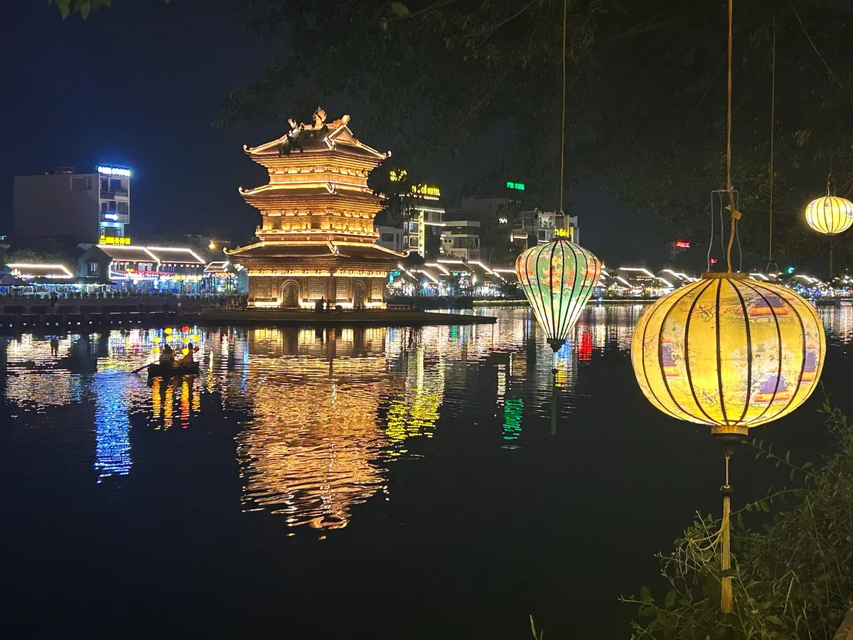 Illuminated pagoda reflects on a river at night, with colorful hanging lanterns and a boat carrying lit balloons. Urban skyline in background.
