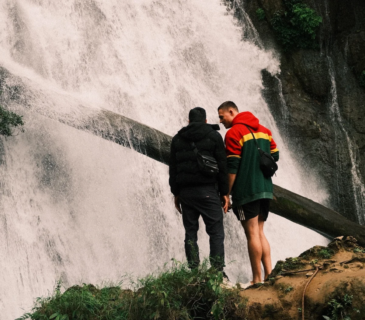 Two people stand on a rocky ledge in front of a waterfall. One wears a colorful hoodie. The scene is natural and dynamic, with cascading water.