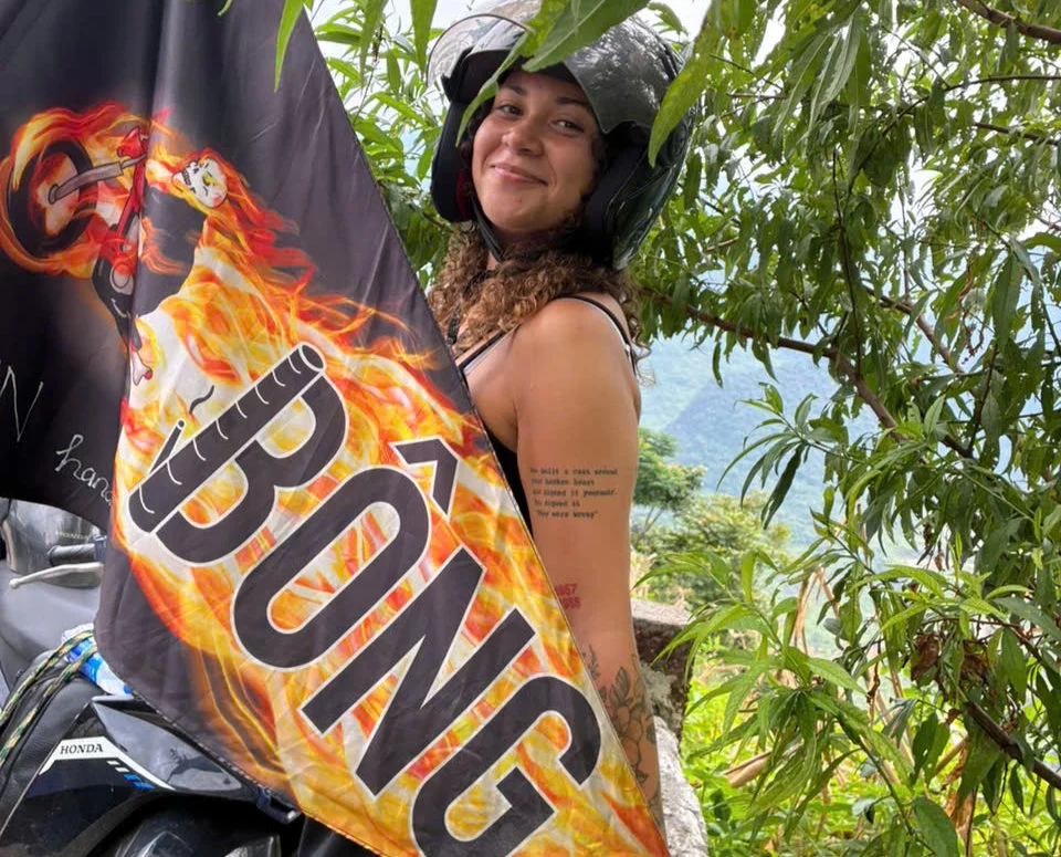 Woman with helmet smiles beside a flaming flag reading "BONG" amid tropical scenery. Text tattoo on arm, lush greenery in background.