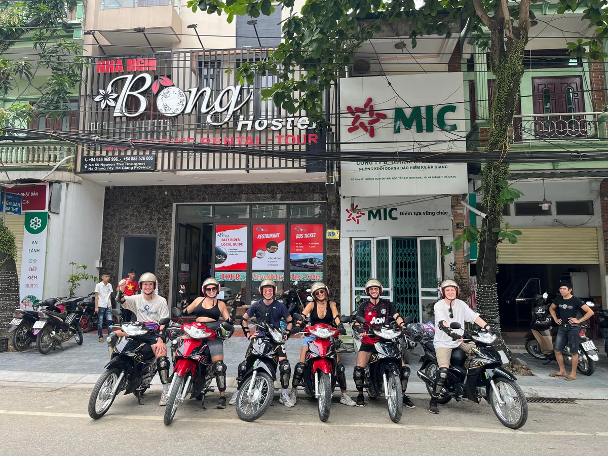 Six people on motorcycles in front of "Bong Hostel" sign, smiling and wearing helmets. Street setting with green and red signs. Energetic mood.