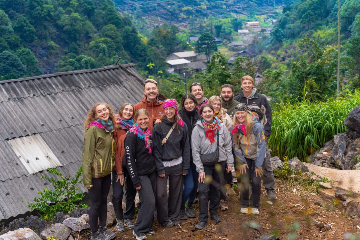 Group of smiling people in colorful scarves pose outdoors on a hillside with lush greenery and distant village, conveying a joyful mood.