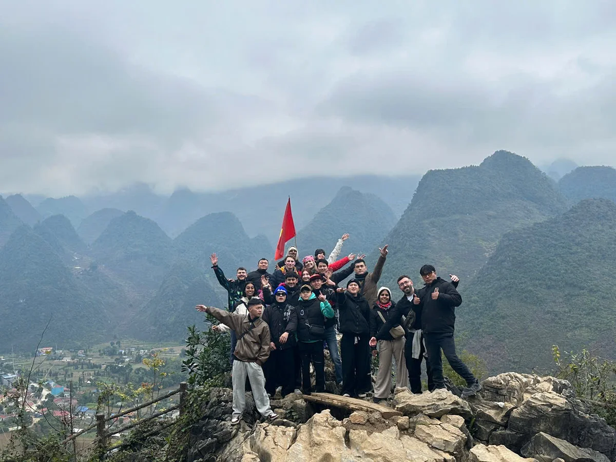 Group of people cheerfully posing on a rocky hilltop, waving a red flag, with misty mountains in the background on a cloudy day.