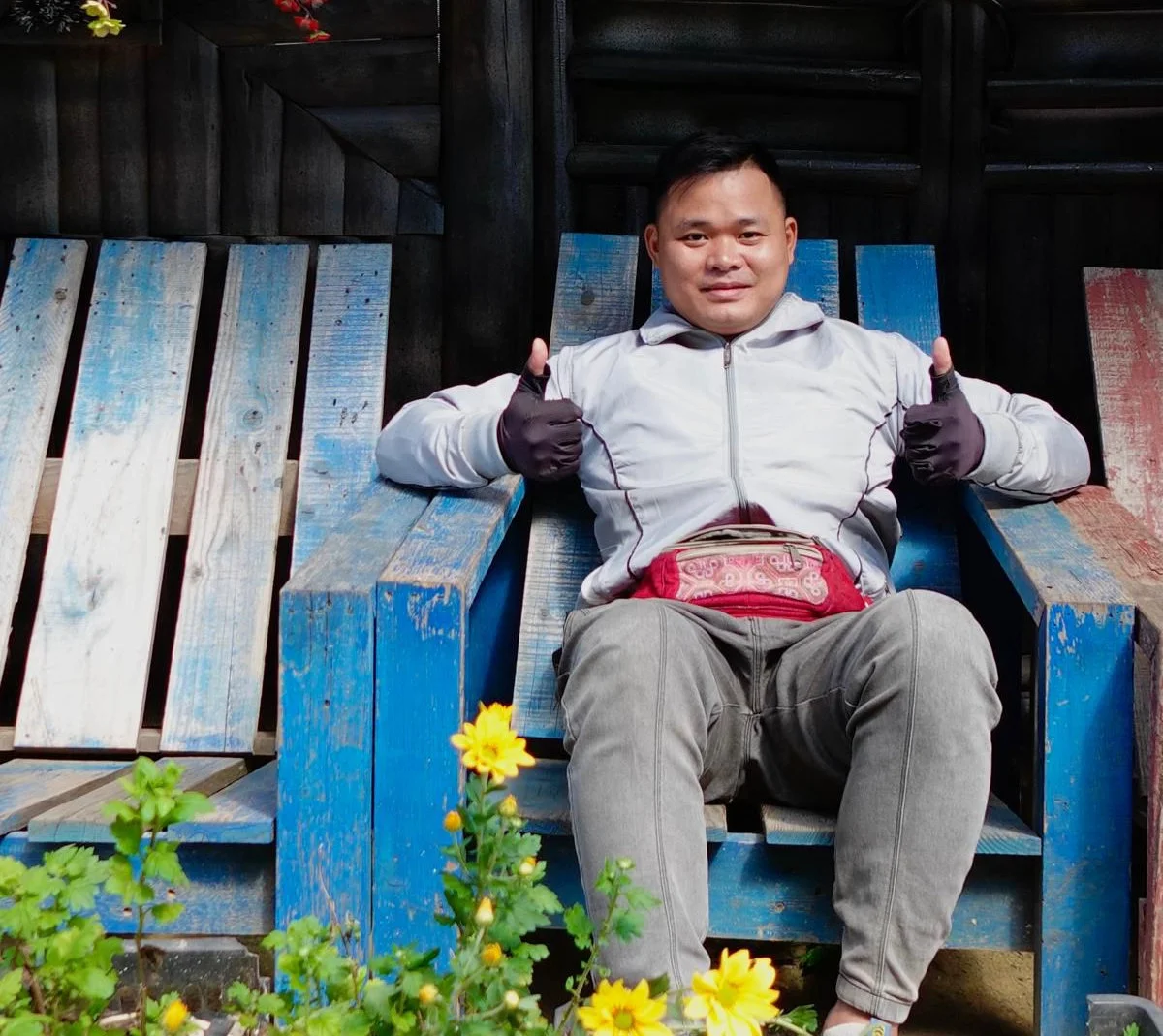 Man in a white jacket and gloves giving thumbs up, seated on a blue wooden bench. Yellow flowers in the foreground, creating a cheerful mood.