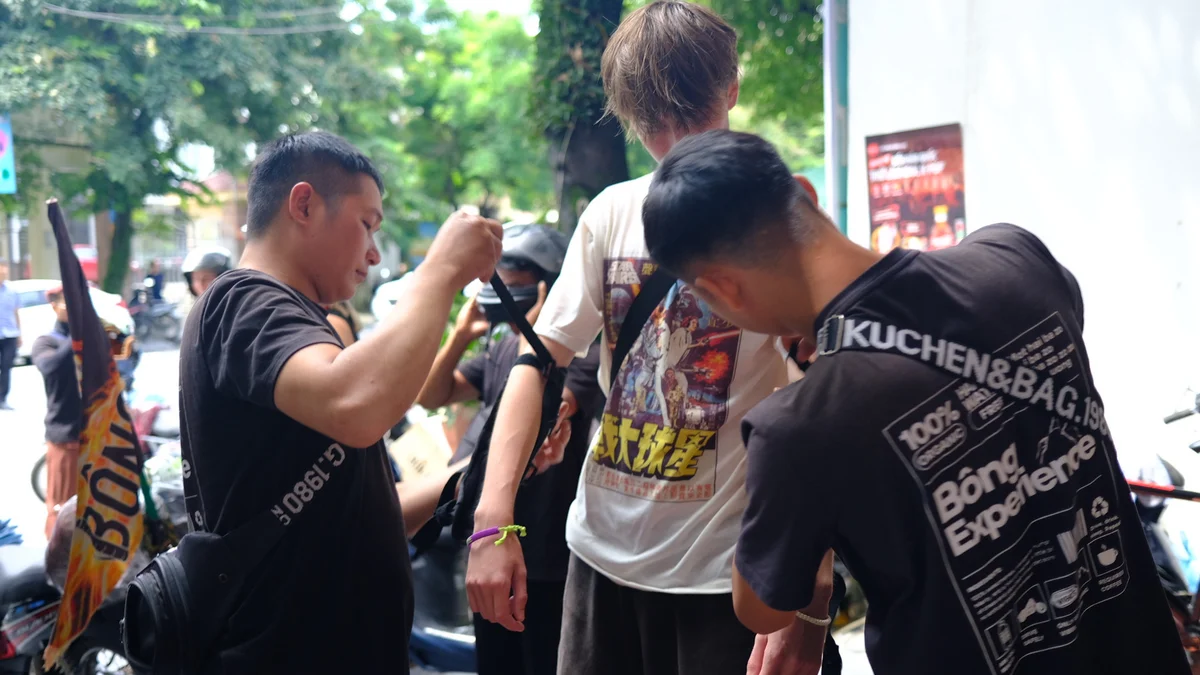 Three people adjusting a backpack outdoors. One wears a graphic t-shirt; another has "KUCHEN&BAG" on their shirt. Trees and motorbikes nearby.