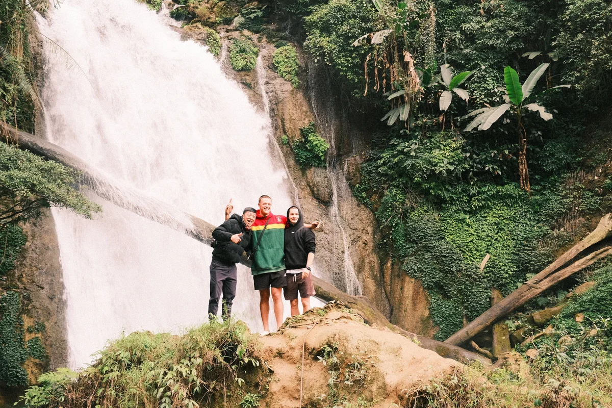 Three people pose happily near a waterfall, surrounded by lush greenery. They're smiling, wearing casual clothing. The scene feels vibrant and cheerful.
