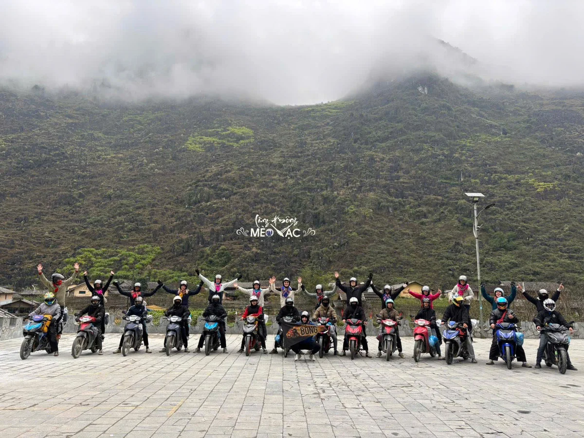 A group of motorcyclists in helmets raise arms joyfully against a foggy mountain backdrop, with text "Mèo Vạc" visible on a sign.