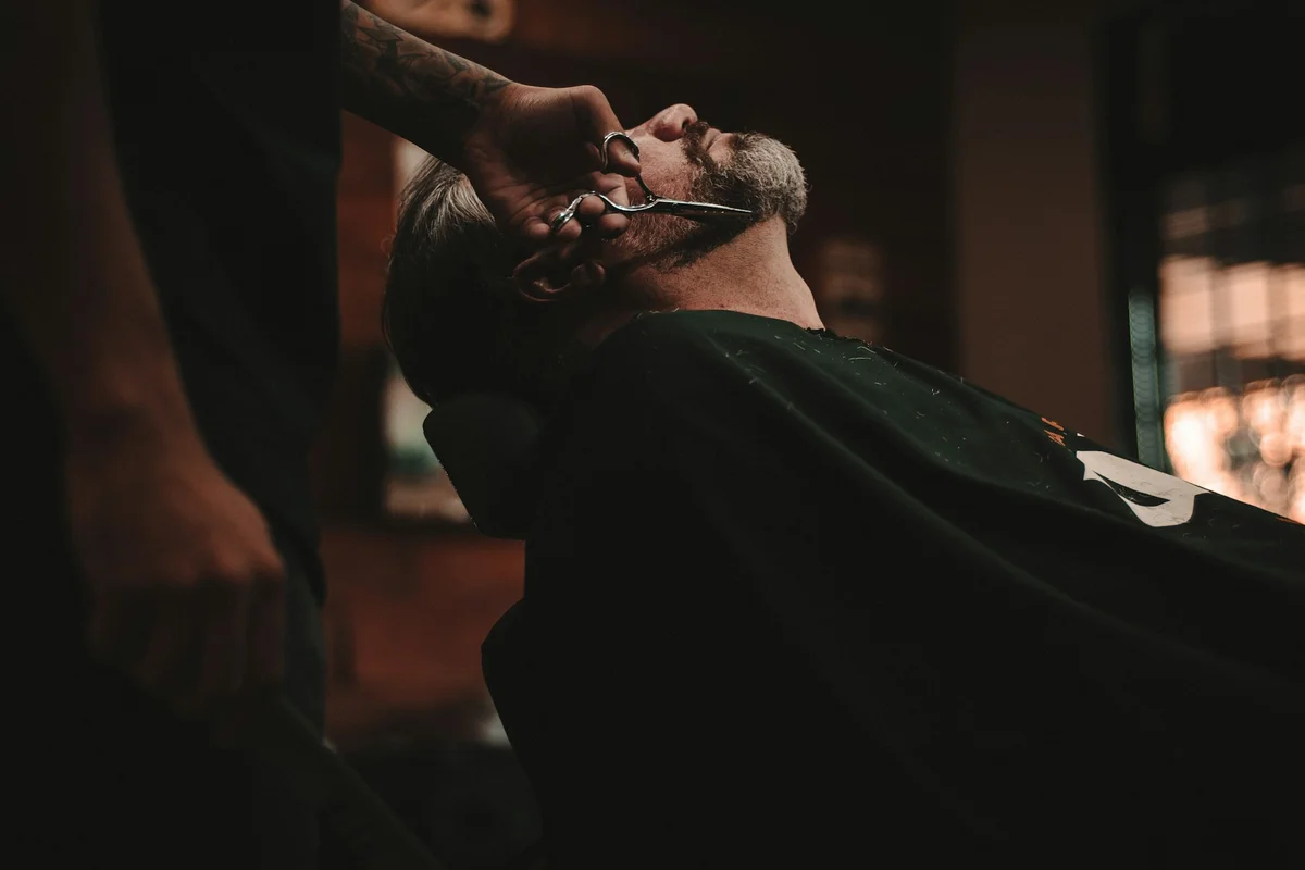 Man leans back in a chair and gets beard trimmed with scissors