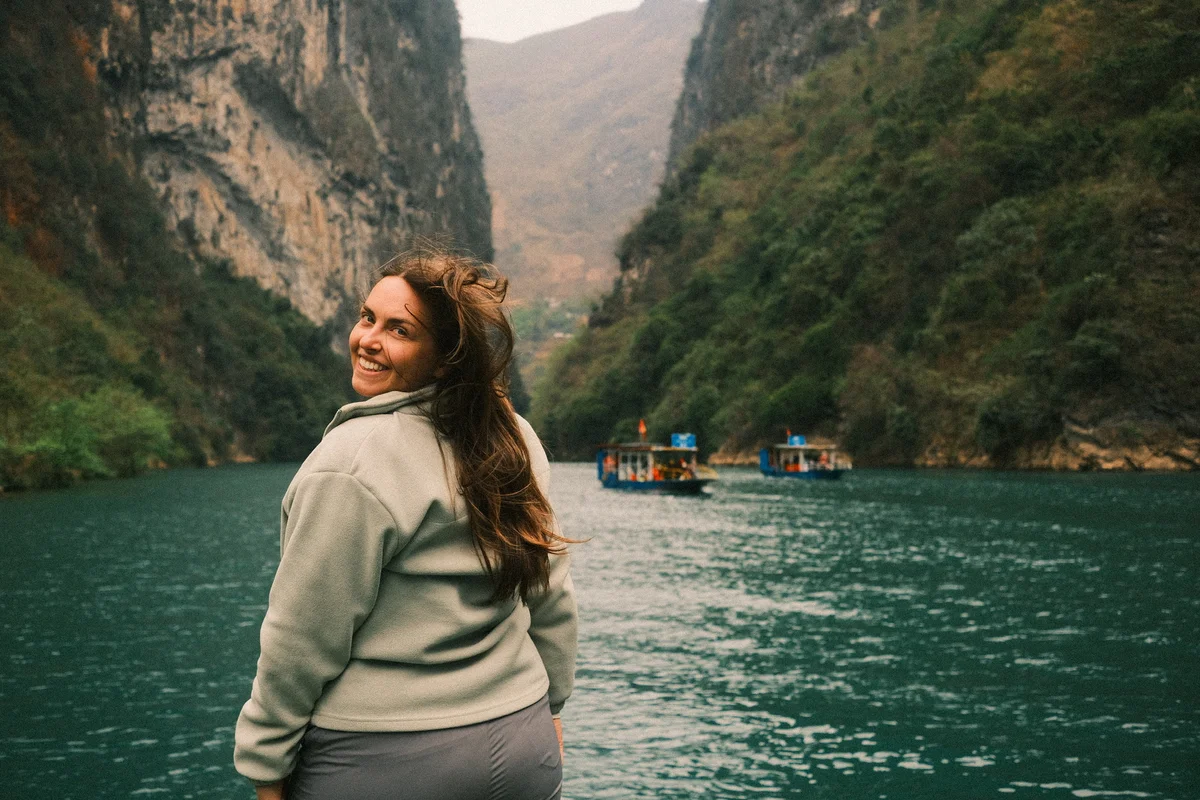 Smiling woman in a gray jacket stands near a turquoise river, flanked by tall cliffs. Boats are visible in the background. The mood is joyful.