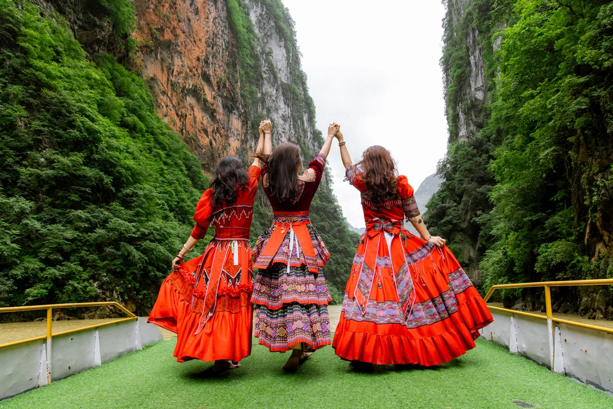 Three women in vibrant red dresses hold hands joyfully on a boat amid lush green cliffs and a clear sky, creating a festive mood.
