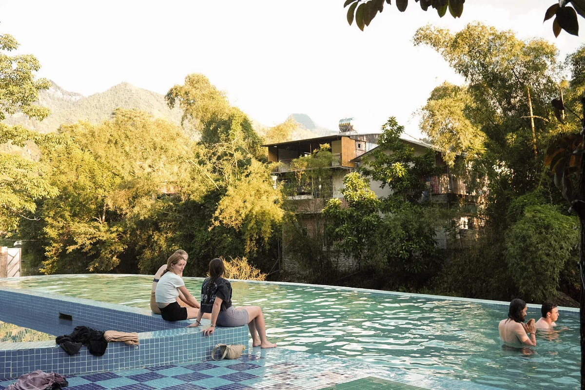 Two people sit on a pool edge, smiling, with lush greenery and buildings in the background. Other people relax in the water nearby.