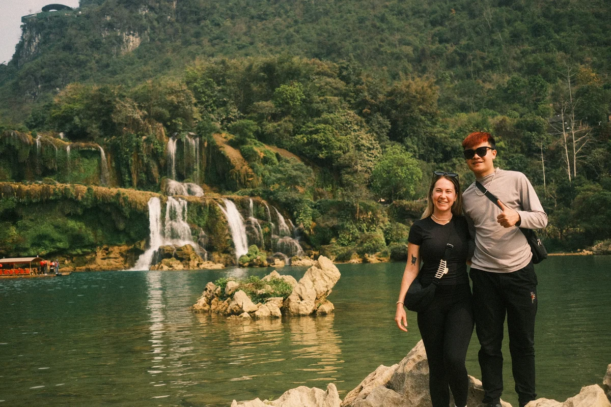 A smiling couple poses by a scenic waterfall, surrounded by lush greenery and a calm lake. One gives a thumbs up, creating a joyful mood.