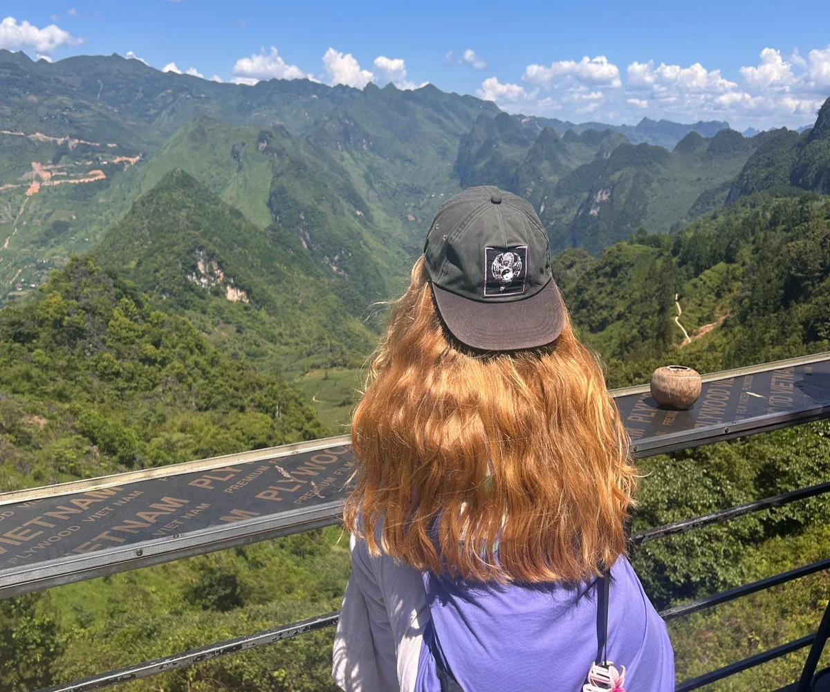A person with long hair and a cap gazes at lush green mountains under a clear blue sky. A railing features "VIETNAM" text. Serene mood.
