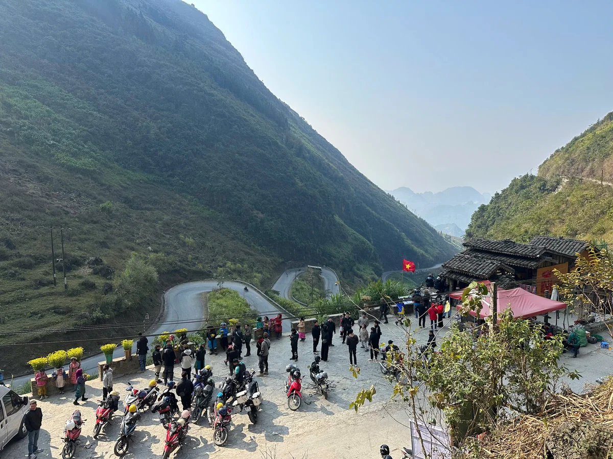 Crowd with motorbikes gathers by a winding mountain road. Bright flags and flowers decorate the area. Mood is lively amidst the lush landscape.