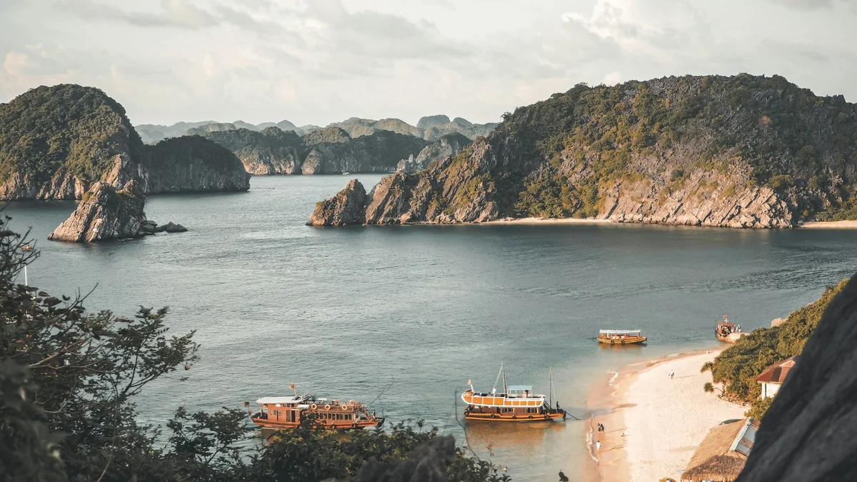 Serene bay with four boats near a sandy beach, bordered by lush green cliffs under a cloudy sky. People are walking on the shore.