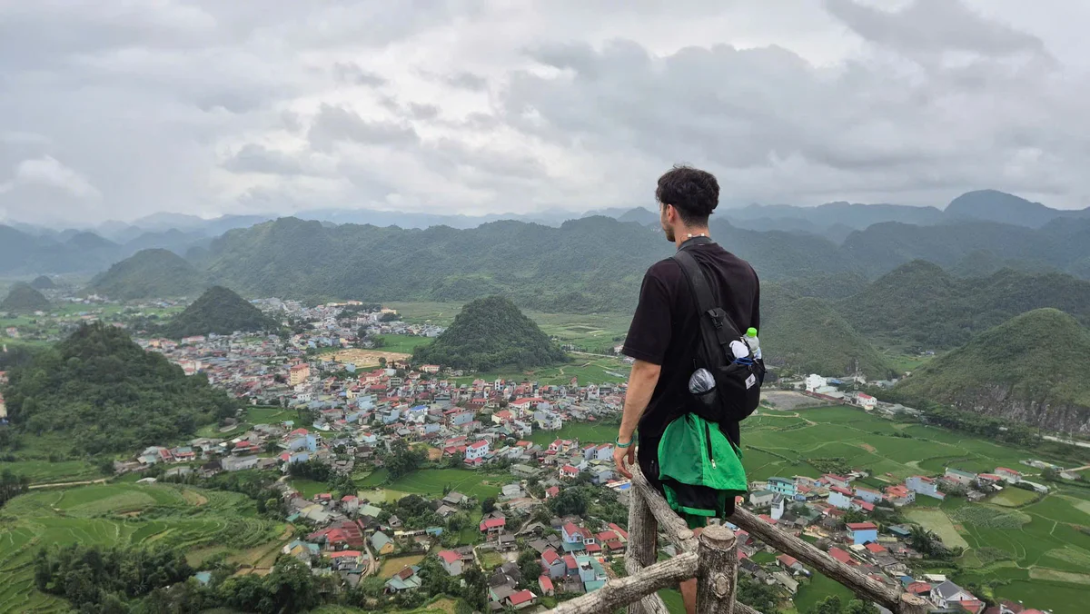 Man with backpack overlooks scenic valley town and rolling green hills under a cloudy sky, evoking a sense of adventure and tranquility.