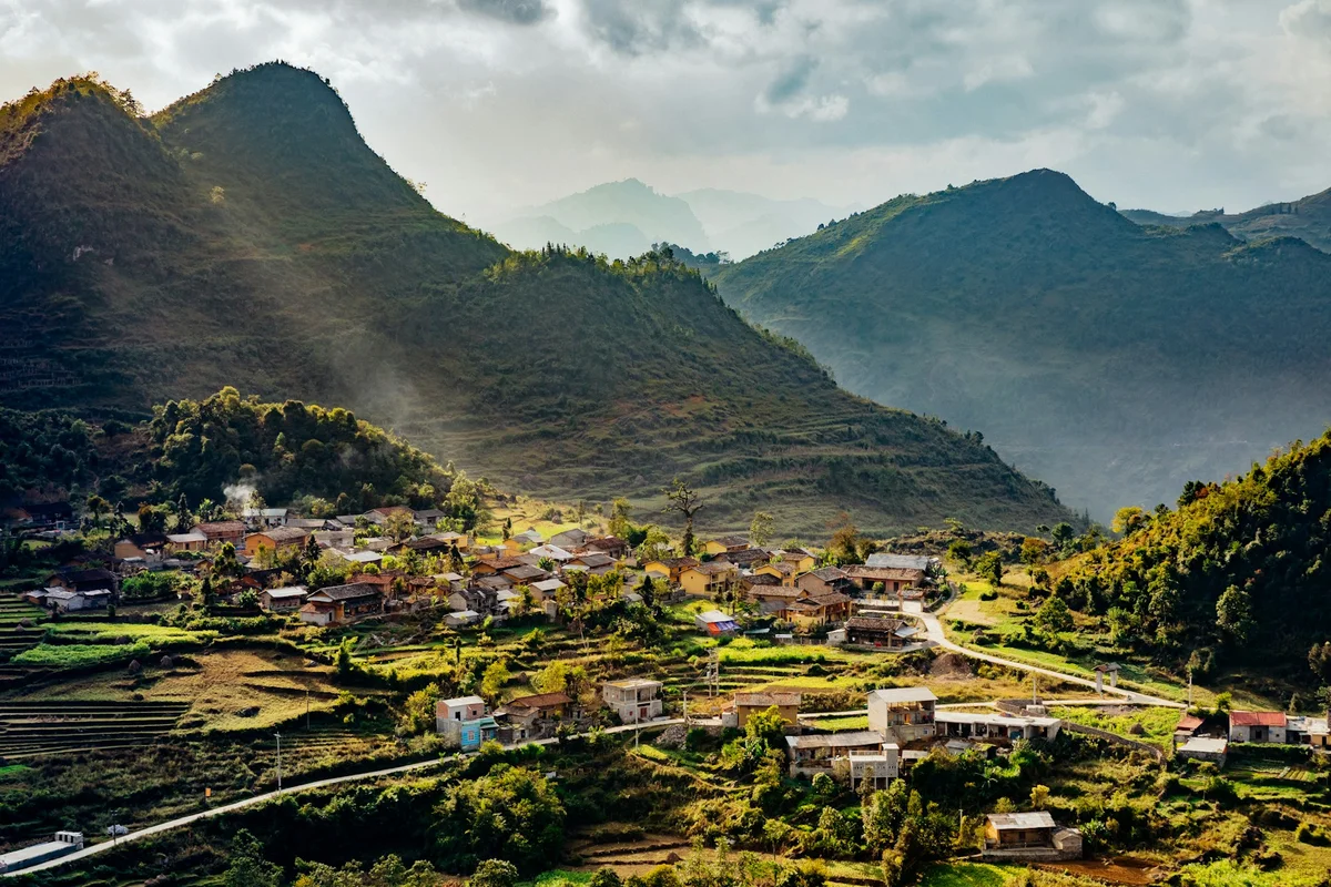 Mountain village landscape with sunlight streaming through clouds, houses nestled among green hills, creating a serene and peaceful scene.