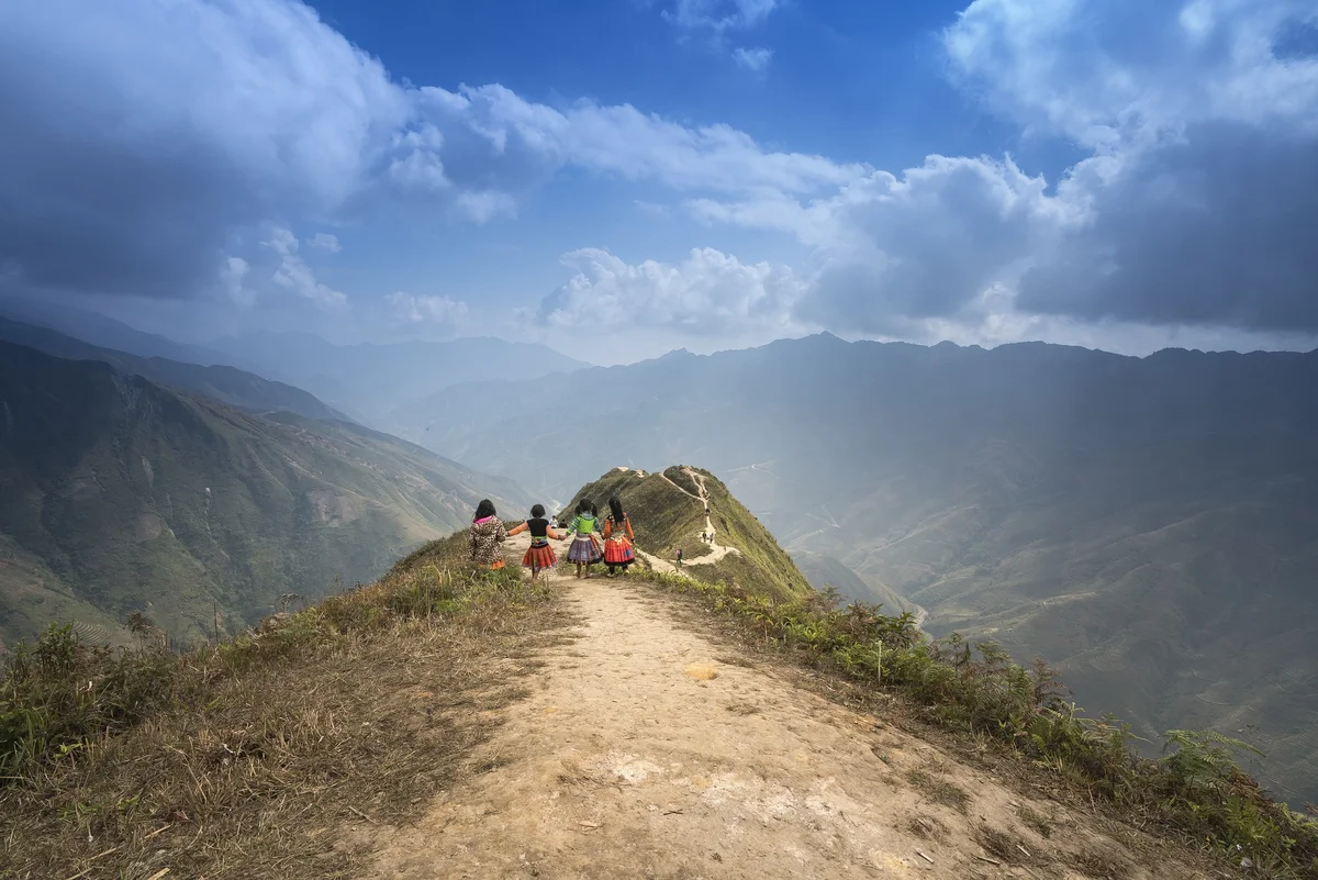 Four children in colorful clothes walk on a mountain path with a stunning view of green peaks and a bright blue sky above.
