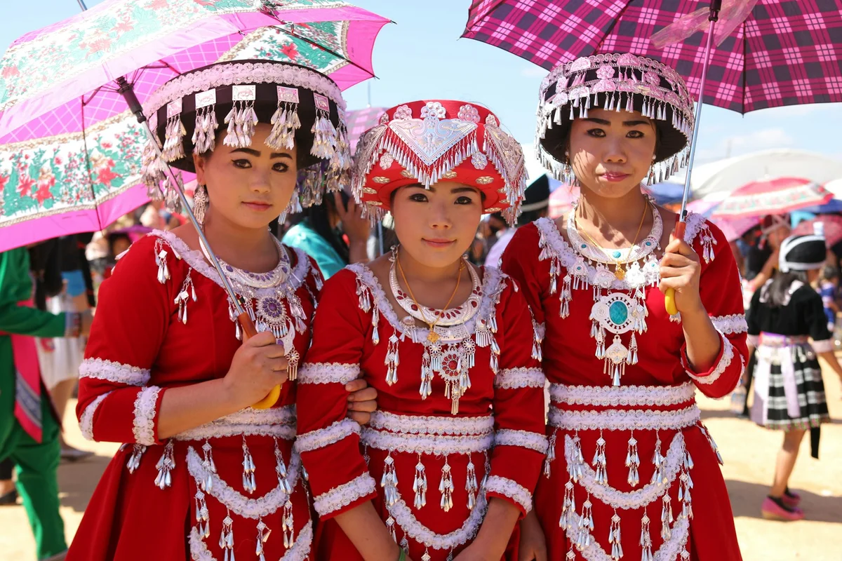 Three people in red traditional attire with silver embellishments hold colorful umbrellas. They stand outdoors on a sunny day, smiling.