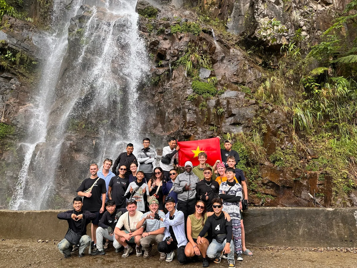 Group poses happily by a waterfall, holding a red flag with a yellow star. Rocky, lush background; casual attire, joyful atmosphere.