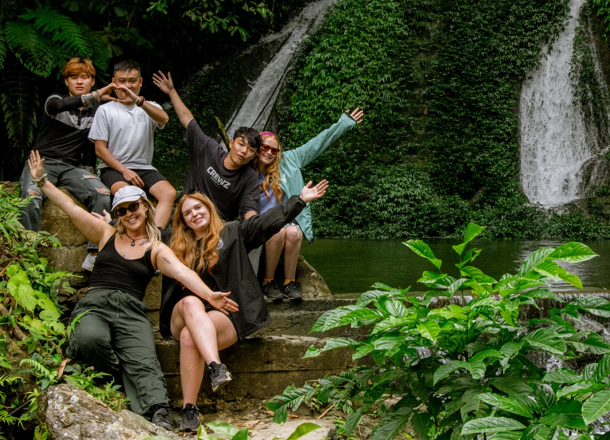 Six people pose happily with arms raised near a waterfall, surrounded by lush greenery. They appear joyful and relaxed.