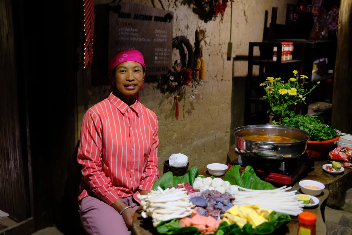 Woman in a pink striped shirt and bandana smiles beside a table of vegetables and hot pot ingredients. Rustic decor, leafy plants, warm lighting.