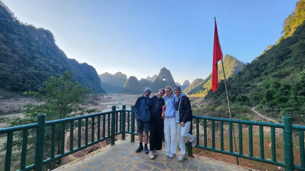 Four people smiling on a viewing platform with stunning mountain scenery in the background. There's a red flag to the right. Bright, clear day.