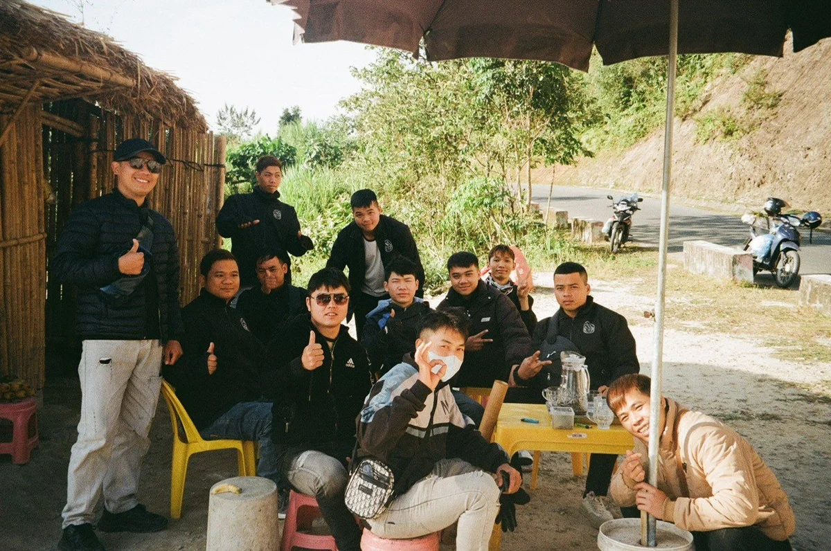 Group of people sitting and standing under a thatched shelter, smiling and giving thumbs up. Road and motorcycles in the background.