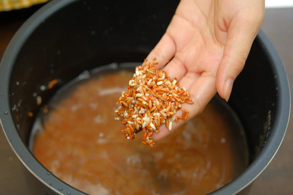 Hand holding a mixture of colorful grains over a pot of water, preparing to cook. The background is a kitchen setting.