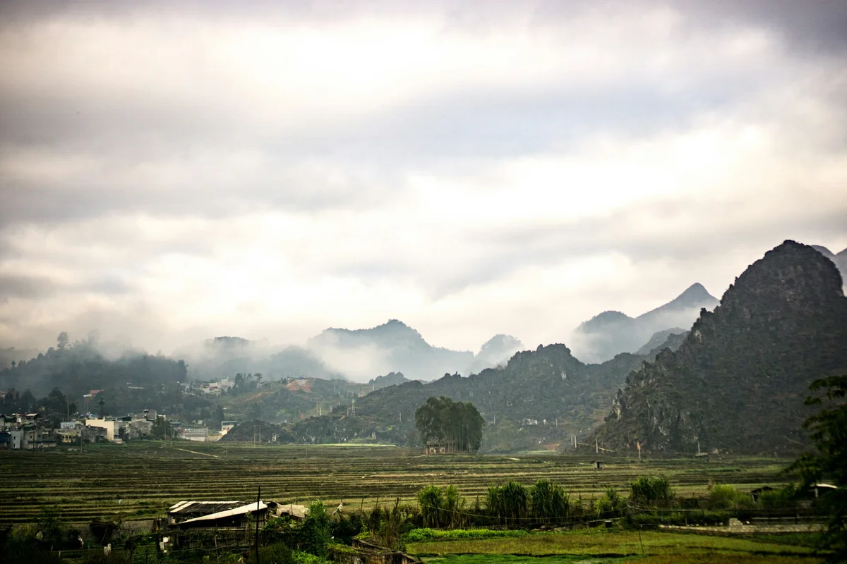 Fog-covered mountains and lush green fields under a cloudy sky create a serene, misty landscape with a village in the distance.