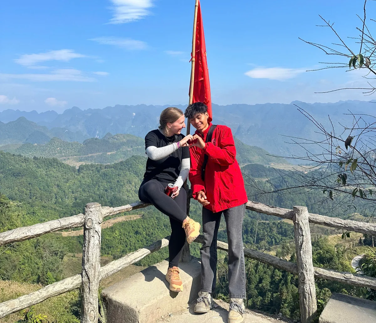 Two people making a heart gesture, one in black, the other in red, on a wooden railing with mountains in the background, a red flag above them.