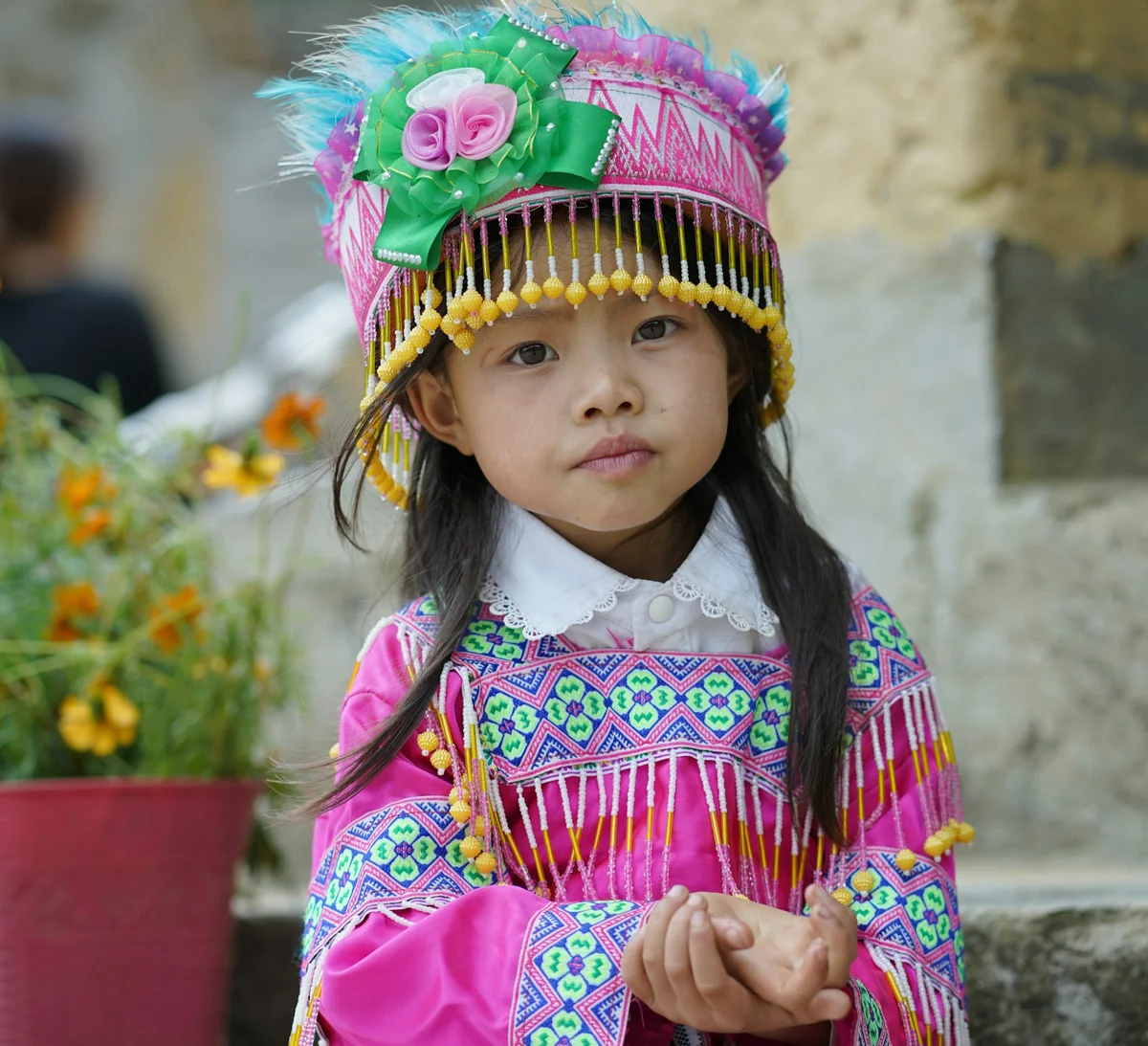 Young girl in traditional pink attire with intricate patterns and a colorful headdress, stands near orange flowers, looking thoughtfully.