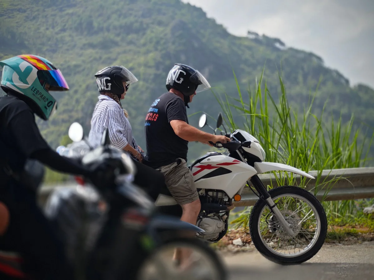 Motorcyclists with helmets ride on a scenic road by lush hills. A white bike with red markings leads, creating an adventurous mood.