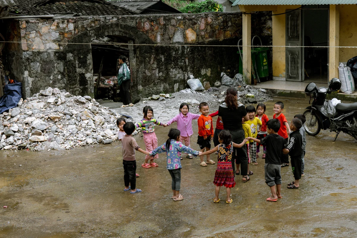Children hold hands in a circle, smiling and playing outdoors. Stone wall and rubble pile in background, motorbike on the right.