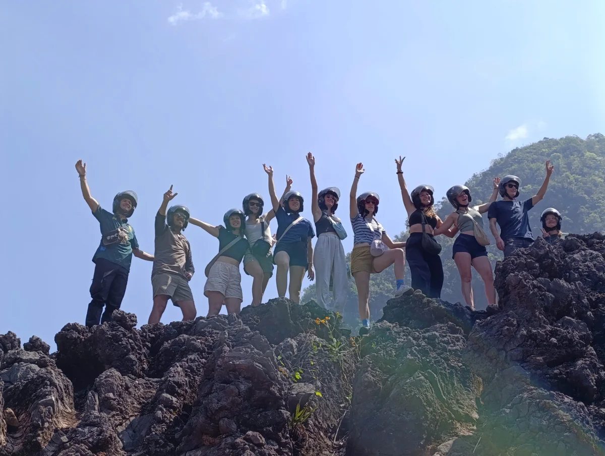 Group of people wearing helmets stand on rocky terrain, raising arms in celebration against a clear blue sky. Mountainous landscape in background.