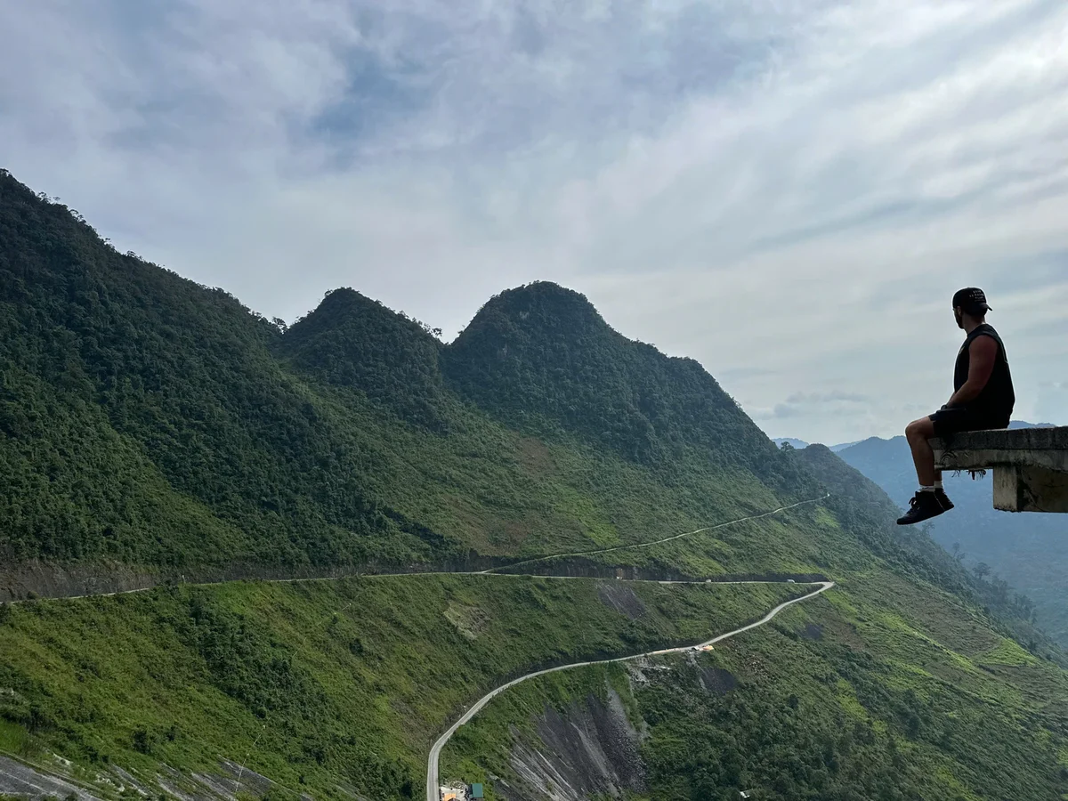 Man in black sits on a ledge overlooking winding mountain road. Lush green hills and cloudy sky create a serene backdrop.