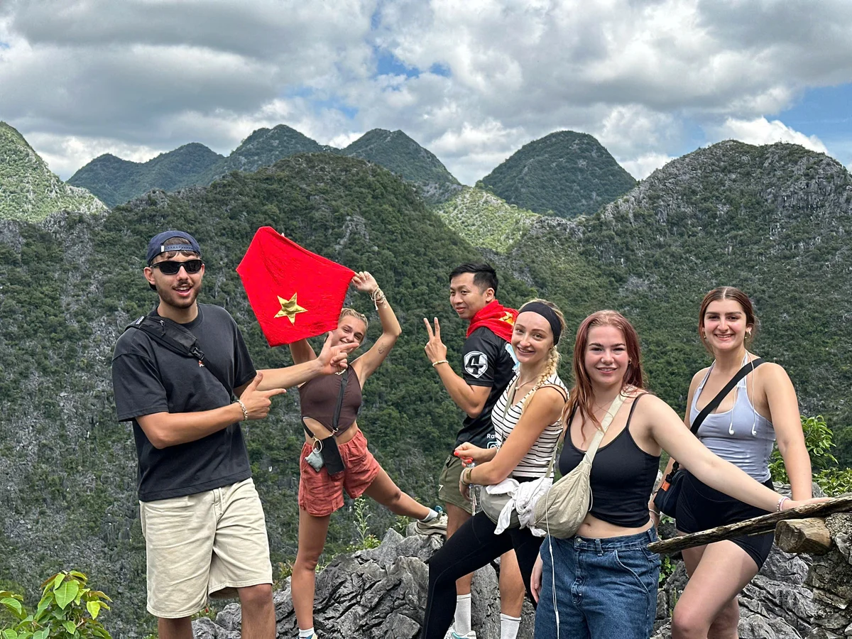 Group of six people posing on a mountain, one holding a red flag with a yellow star. Cloudy sky and green mountains in the background.