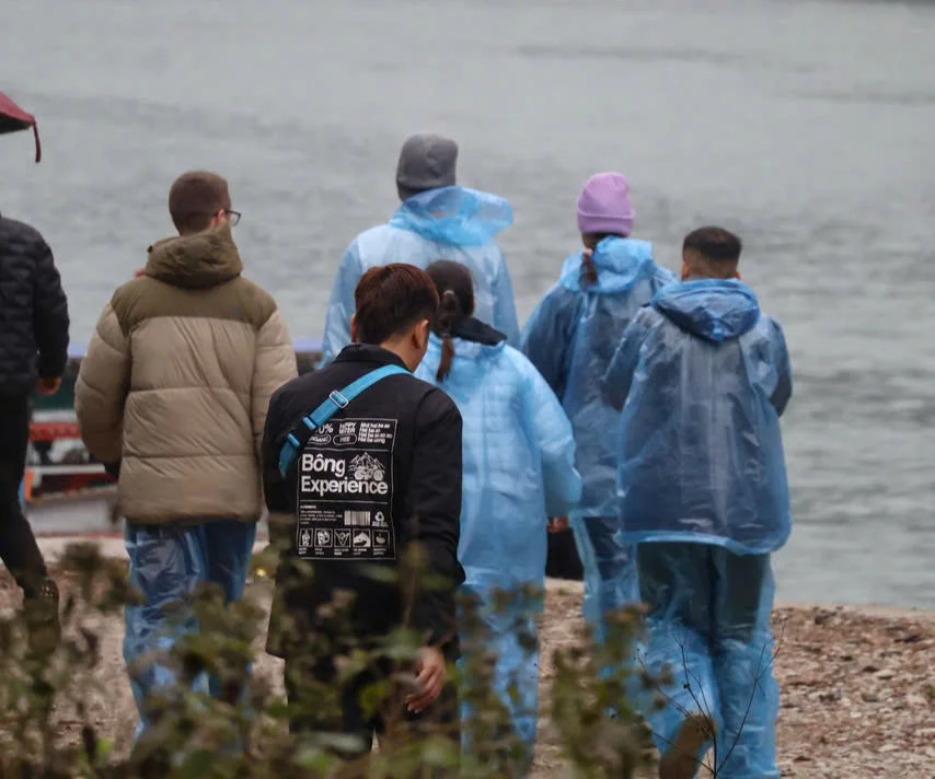 People in blue raincoats walking by a river. One wears a jacket with text "Bong Experience." Overcast setting, muted colors, calm mood.