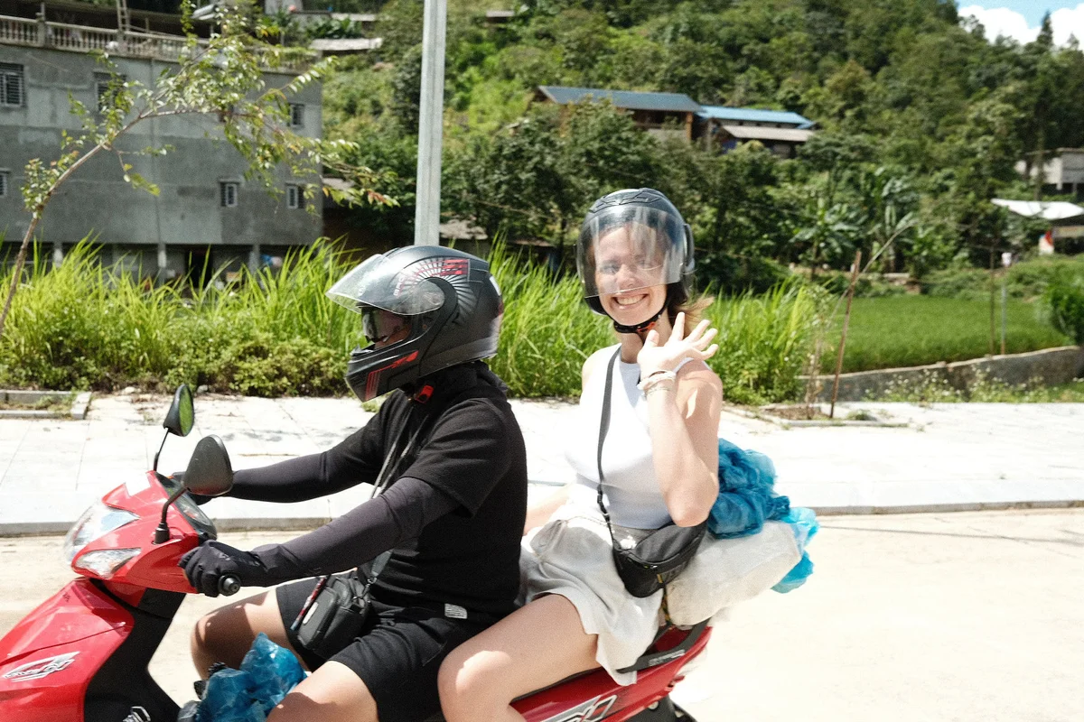 Two people ride a red scooter; one waves happily. They're wearing helmets. Greenery and buildings are visible in the sunny background.