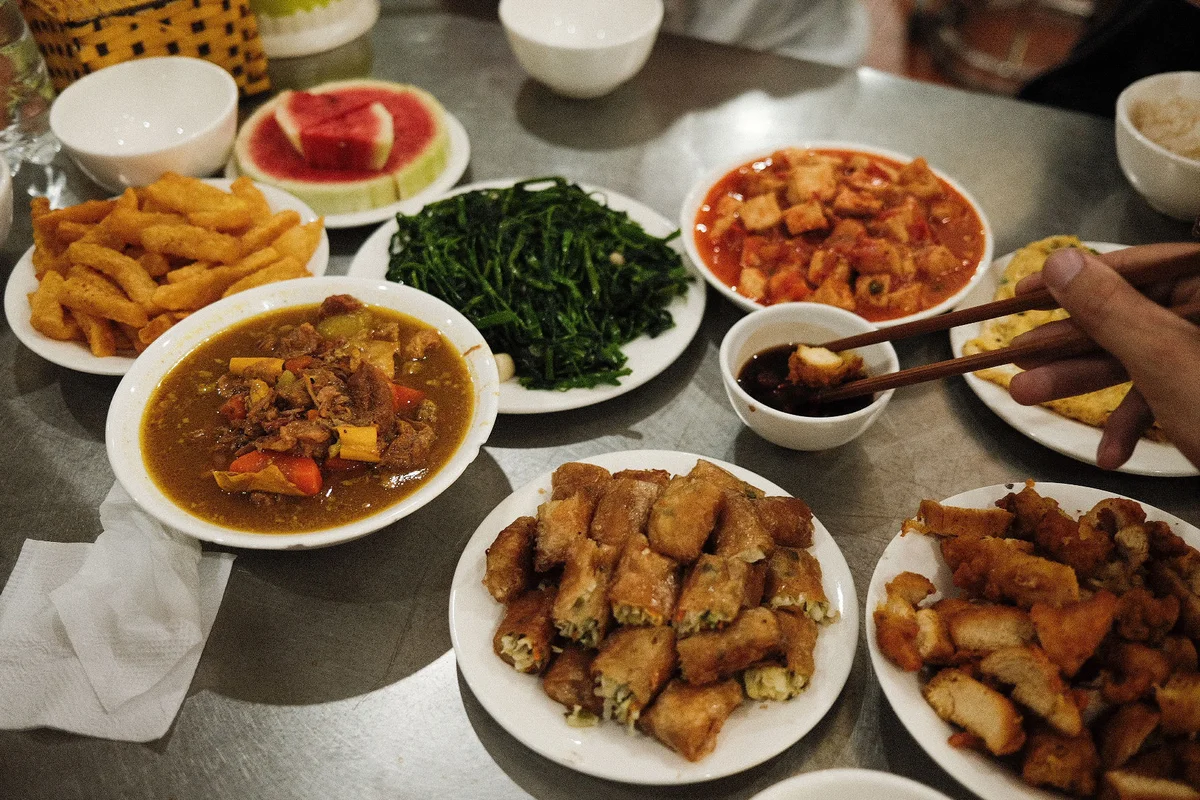 A table with various dishes, including stir-fried greens, curry, and fried rolls. A hand uses chopsticks to dip food in sauce. Vibrant and inviting.