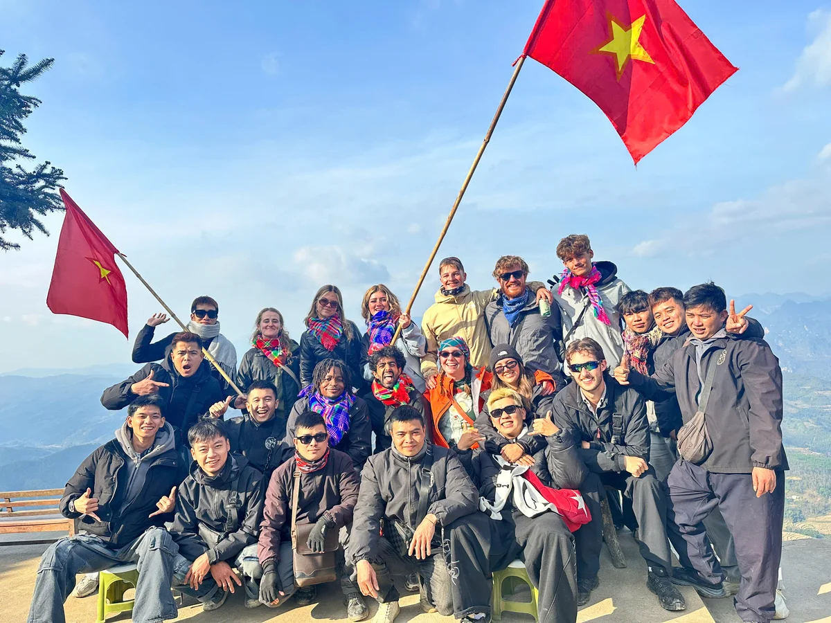 Group of people smiling and posing on a mountain with Vietnamese flags. Bright winter clothing. Clear sky and distant mountains behind.